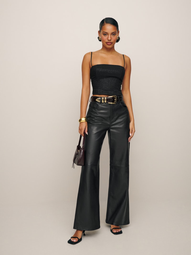 A woman wearing black wide-leg leather pants with a high waist, paired with a black spaghetti strap top, black heels, and gold accessories.
