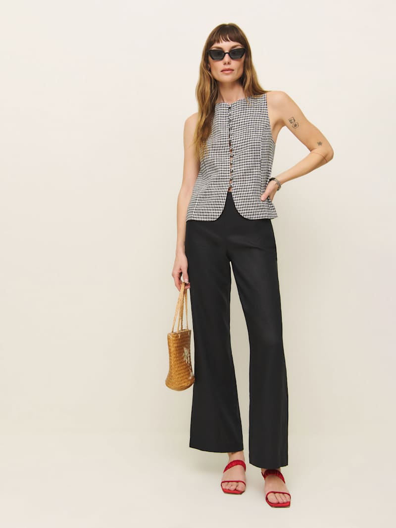 A woman wearing black Petites Hallie Linen Pants, a sleeveless checkered top, red sandals, and sunglasses, holding a woven bag.
