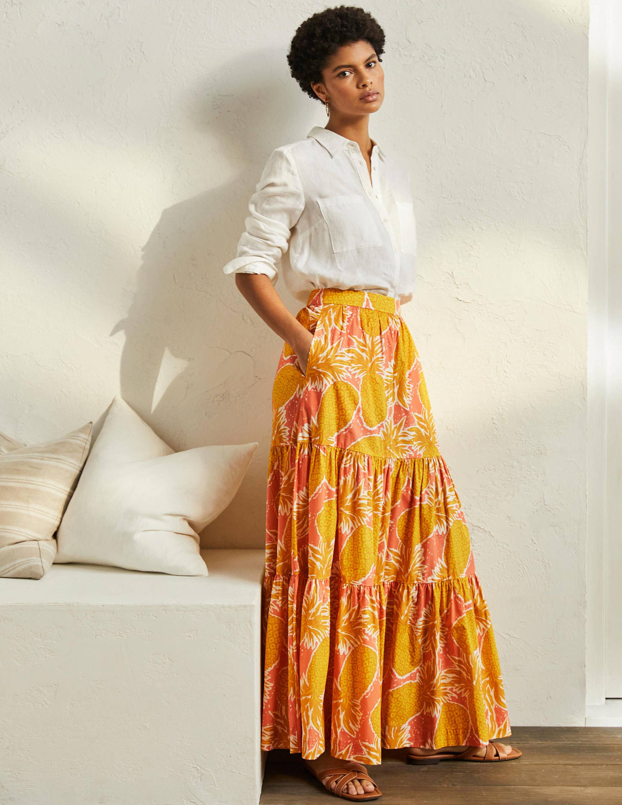 A woman wearing a colorful, tiered maxi skirt with a pineapple and leaf pattern, paired with a white shirt and sandals.