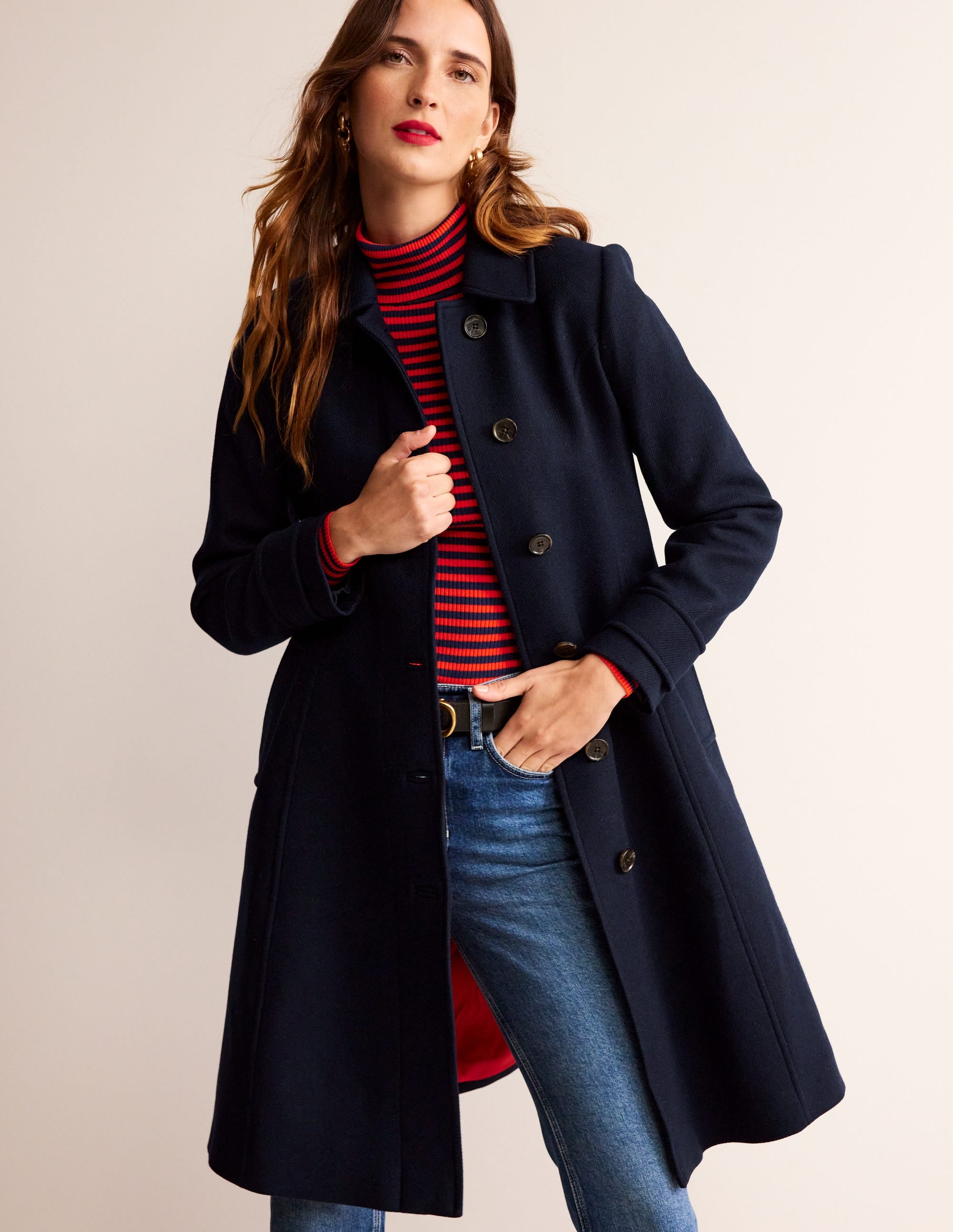 A woman wearing a navy wool blend coat with a tailored fit, princess seams, a smart collar, and button closure, paired with a red and navy striped turtleneck and blue jeans.