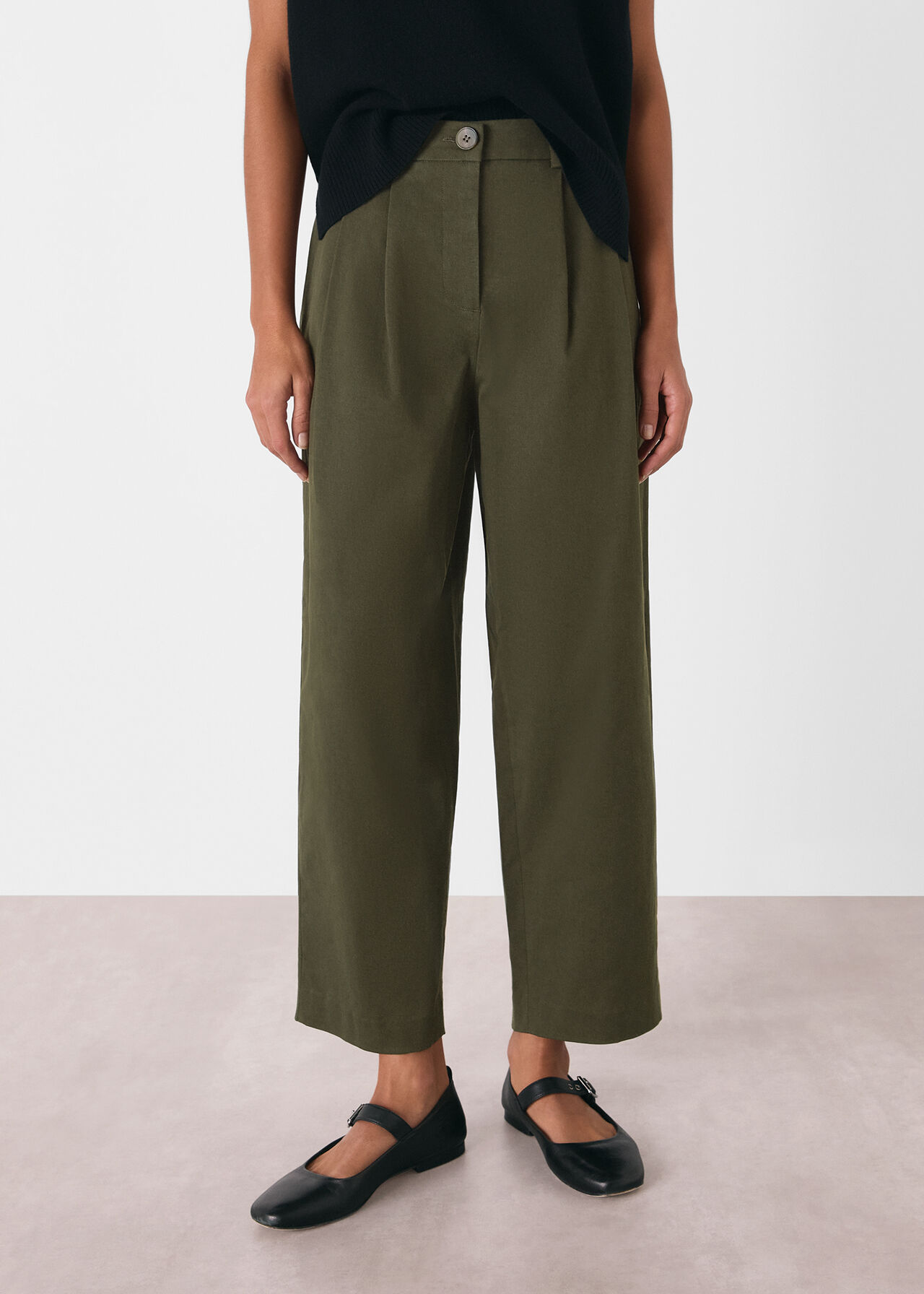 A woman wearing khaki relaxed-fit trousers with a barrel leg shape, two side pockets, and a front button, paired with a black top and black shoes.