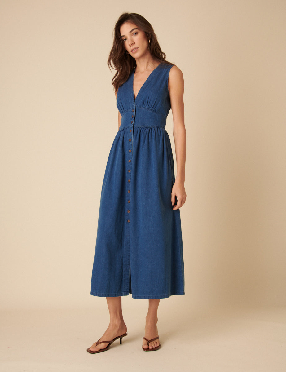 A woman wearing a blue sleeveless denim dress with a V-neckline, button-down front, and ankle-length A-line skirt.