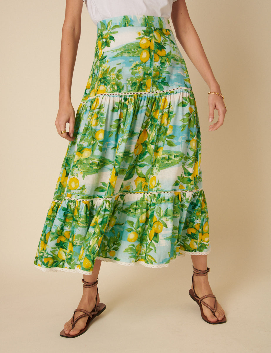 A woman wearing a green linen-blend midi skirt with lemon print, tiered design, and lace trim at the hem.
