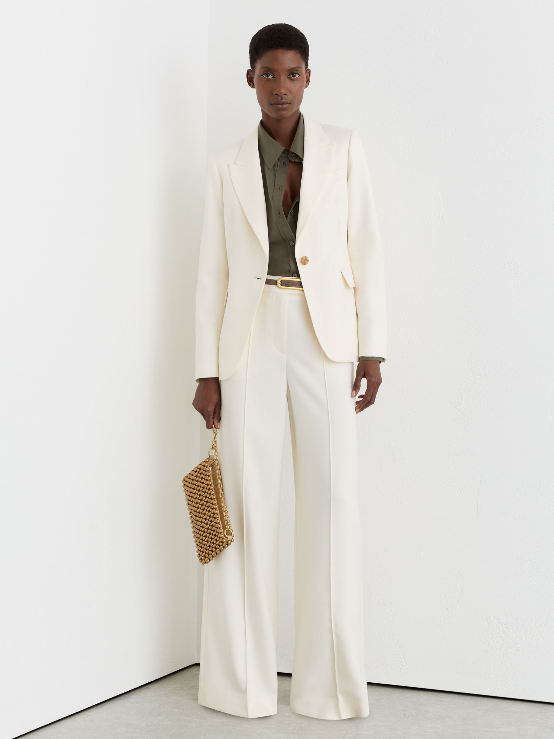 A woman wearing cream wide-leg trousers with front pintucks, a matching blazer, and a dark shirt underneath, holding a gold beaded clutch.