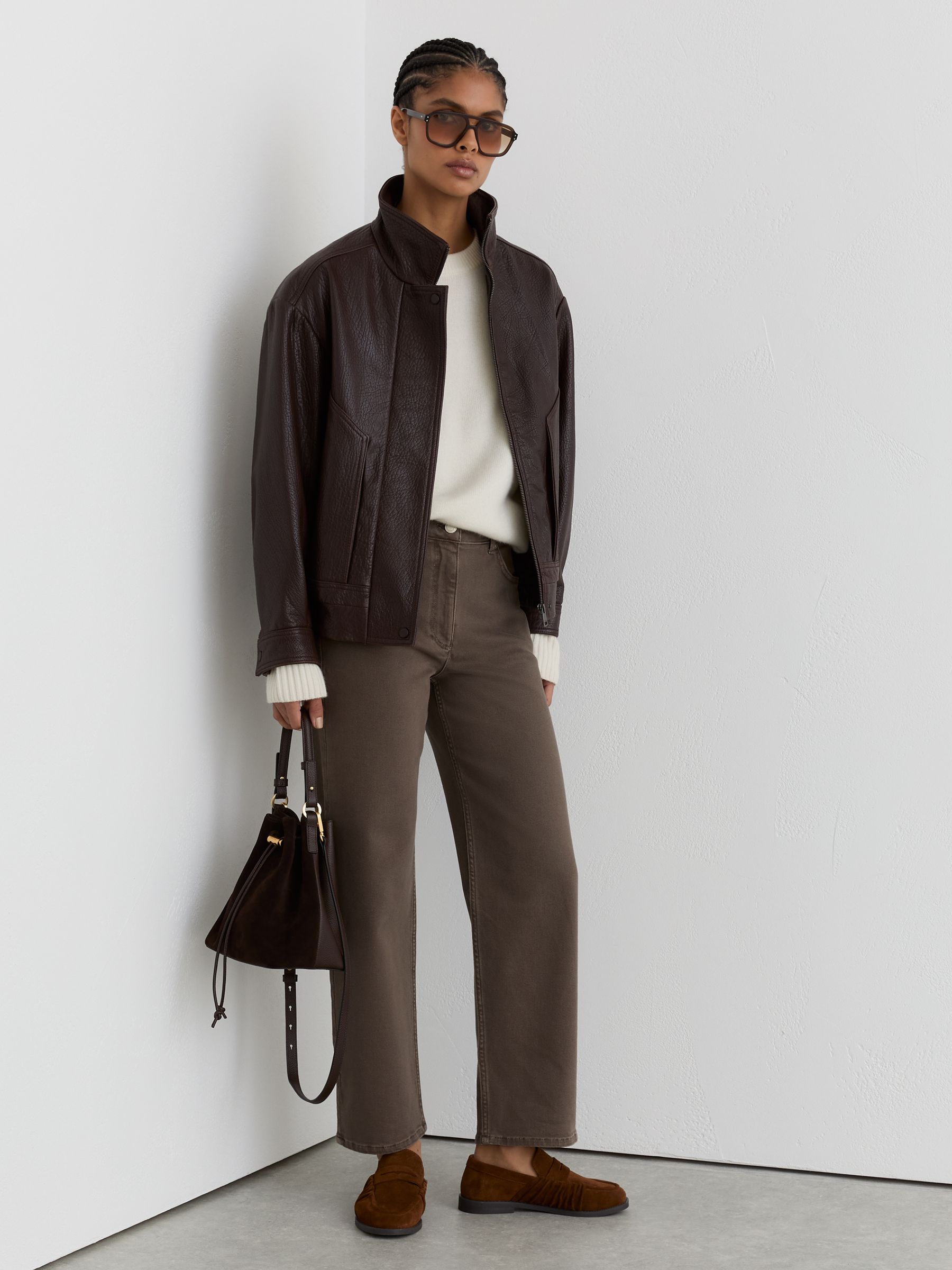 A woman wearing mocha brown straight-leg jeans, a cream sweater, a dark brown leather jacket, brown shoes, and carrying a dark brown handbag, standing against a white wall.