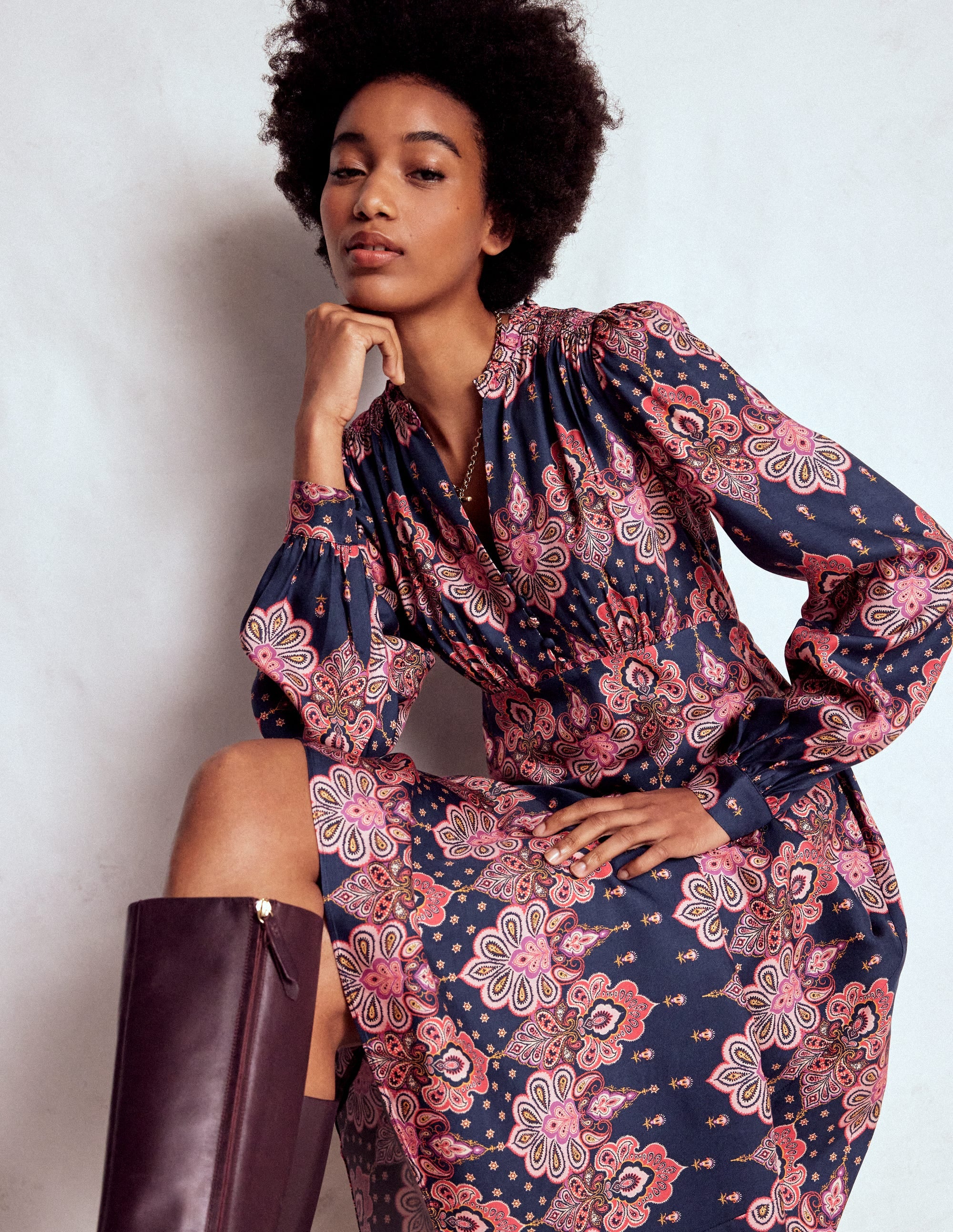 A woman wearing a navy blue smocked tea dress with a pink, white, and gold paisley print, featuring a fit and flare silhouette, in-seam pockets, and a maxi length.