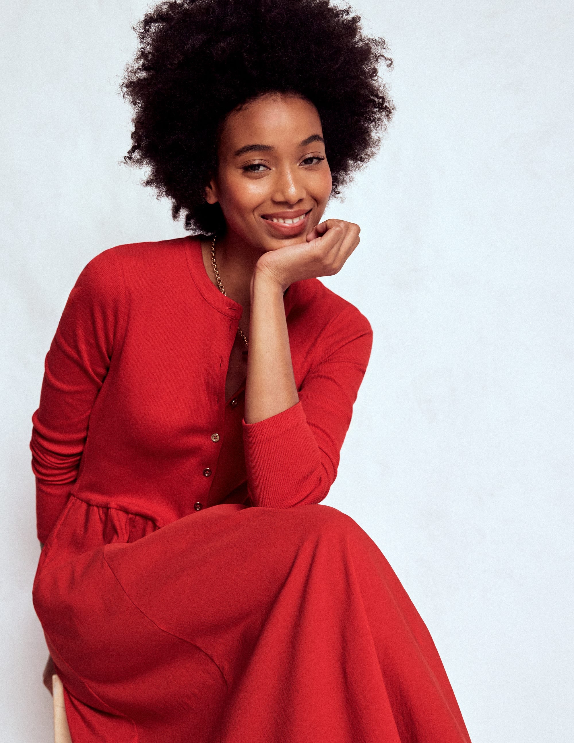 A woman wearing a crimson red dress with a ribbed top and woven skirt, smiling and resting her chin on her hand.