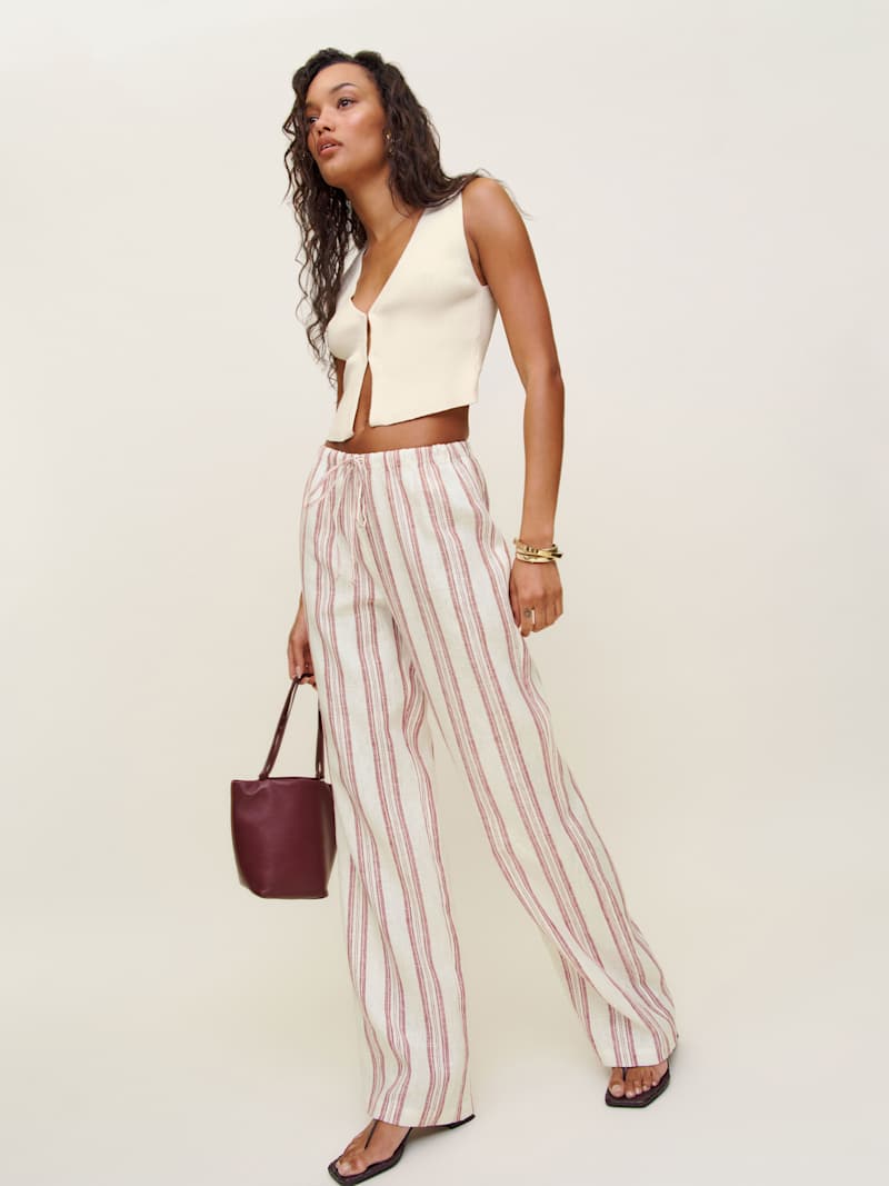 A woman wearing beige linen pants with red and white vertical stripes, a sleeveless cream top, and black sandals, holding a burgundy handbag.