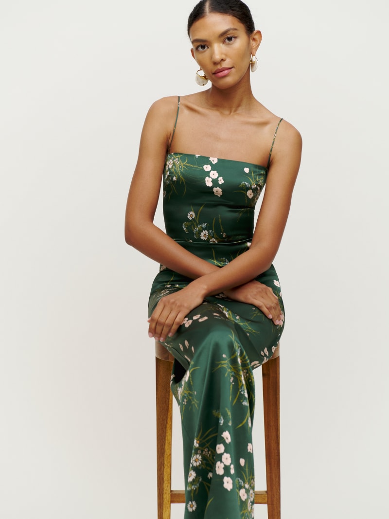 A woman wearing a green floral silk dress with spaghetti straps, a straight neckline, and a center back slit, sitting on a wooden stool.