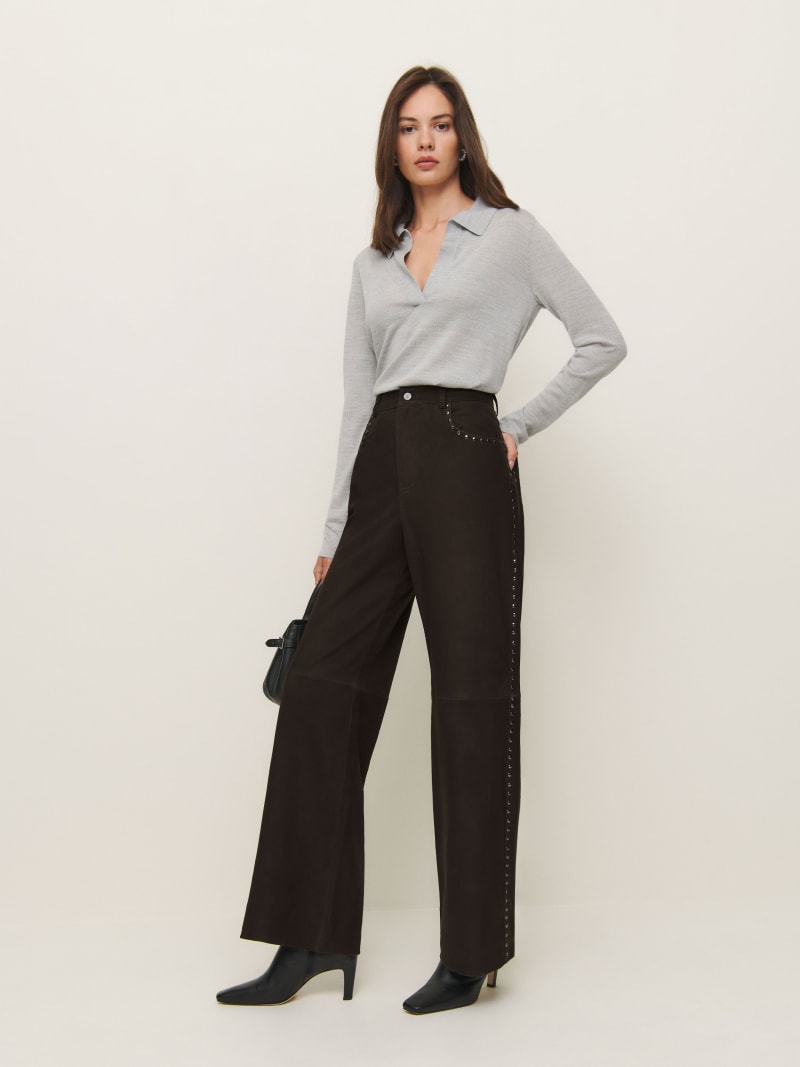 A woman wearing high-rise black leather pants with studded detailing on the side seams, paired with a light gray long-sleeve top and black heeled boots.