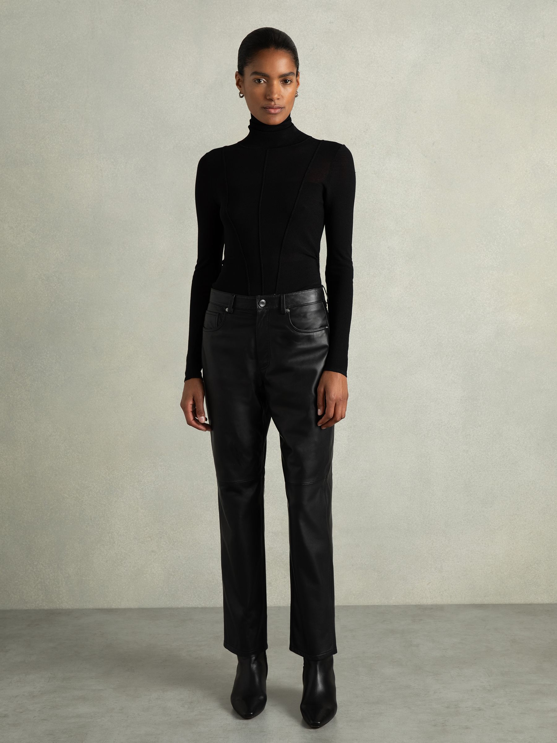 A woman wearing black leather slim-leg trousers with a high waist, belt loops, and five pockets, paired with a black turtleneck top.