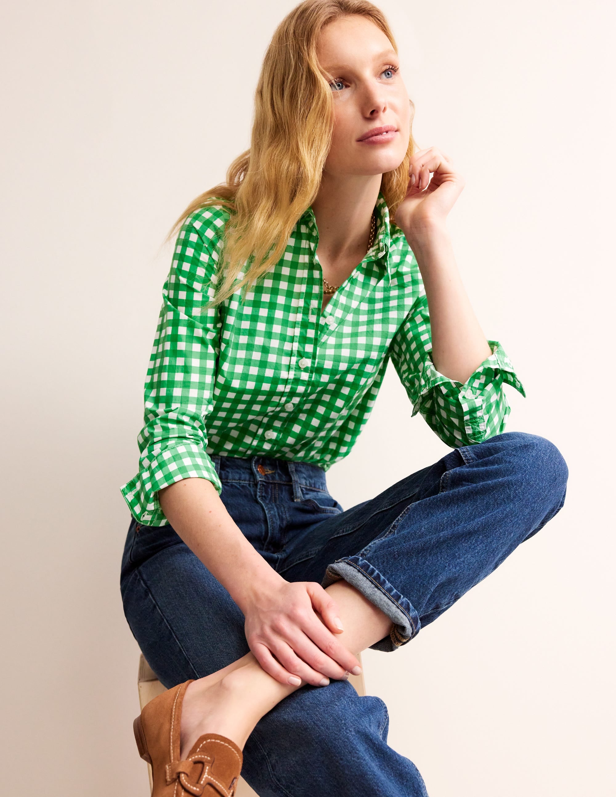 A woman wearing a green gingham cotton shirt with long sleeves and a curved hem, paired with blue jeans and brown shoes.