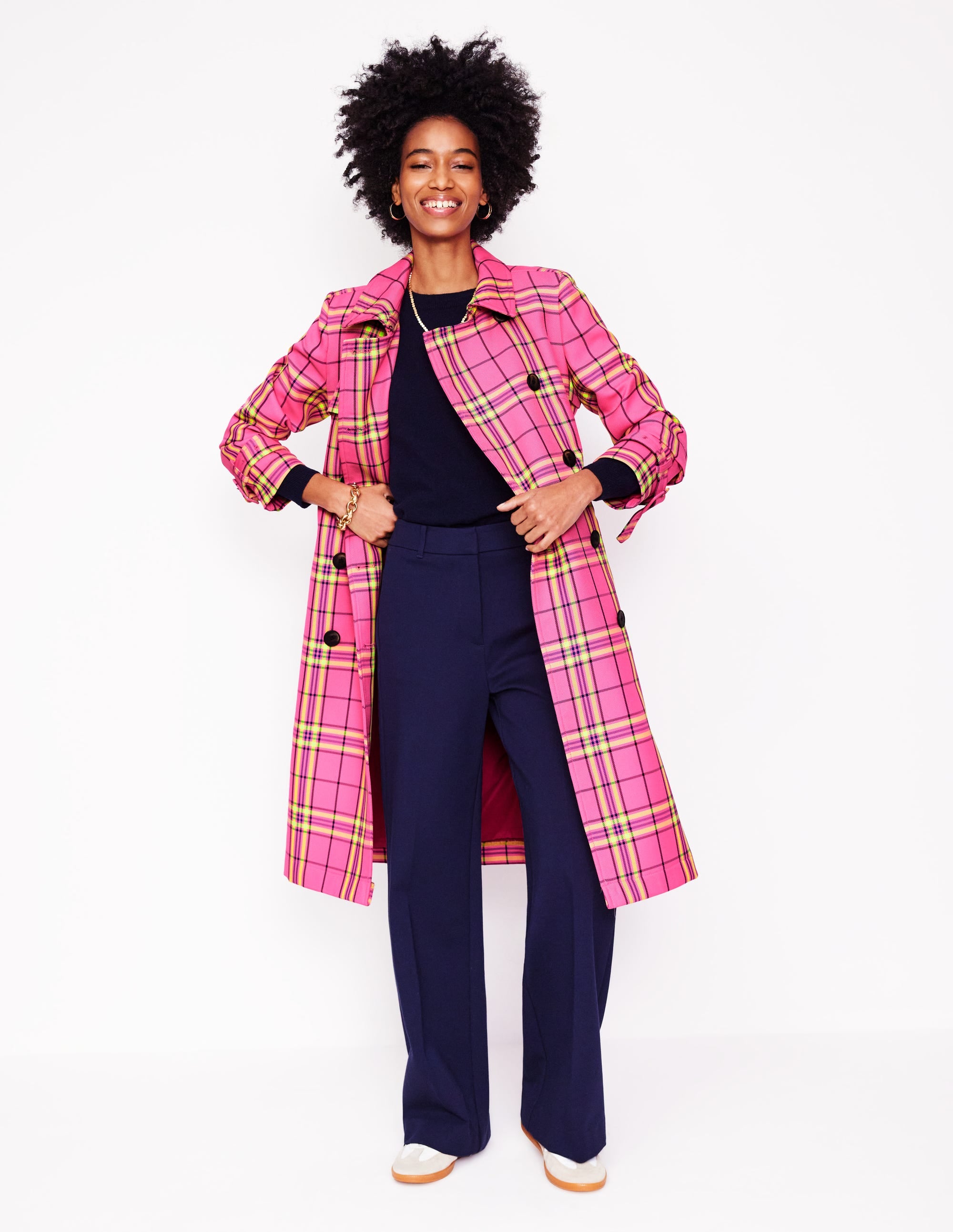A woman wearing a pink neon check trench coat with a double-breasted front, belt, and pockets, standing against a white background.