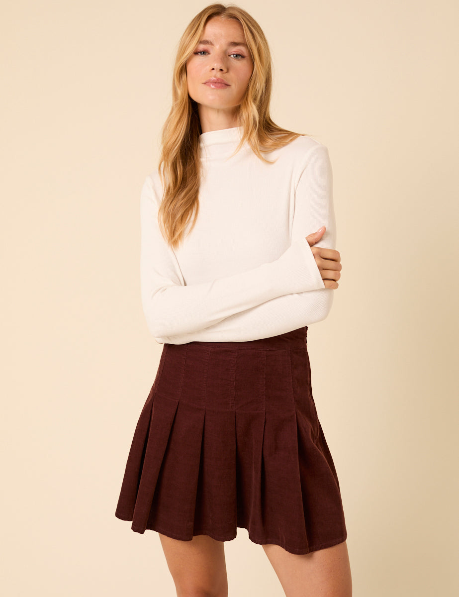 A woman wearing a brown corduroy pleated mini skirt with a high waist, paired with a white long-sleeve top.