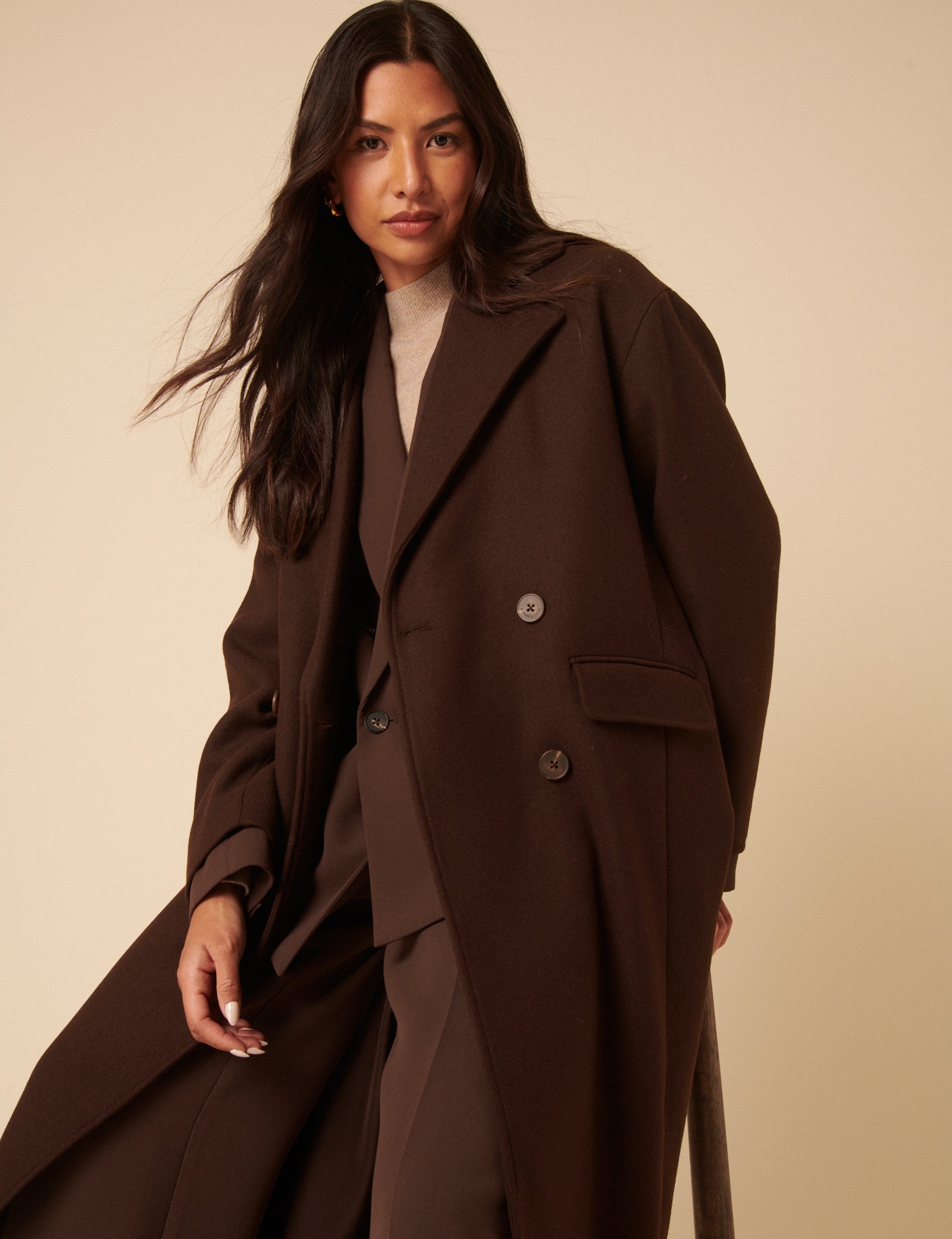 A woman wearing a chocolate brown double-breasted coat with a relaxed fit, featuring a collar, flap pockets, and buttons.