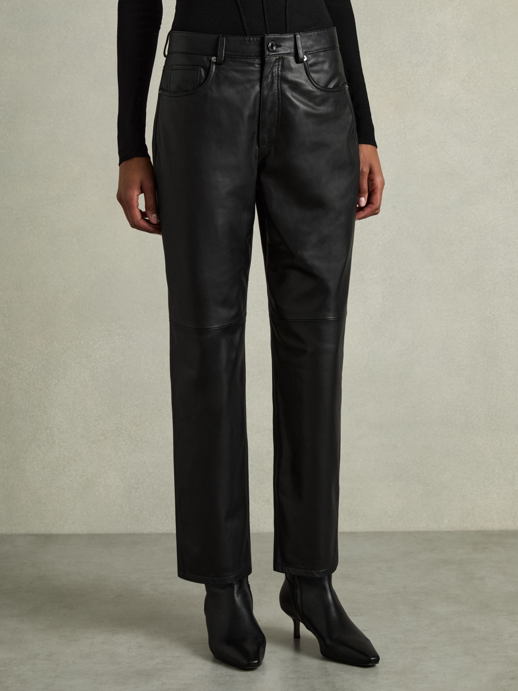 A pair of black leather trousers with a slim-leg design, belt loops, and five pockets, worn by a person with a black top and black boots.